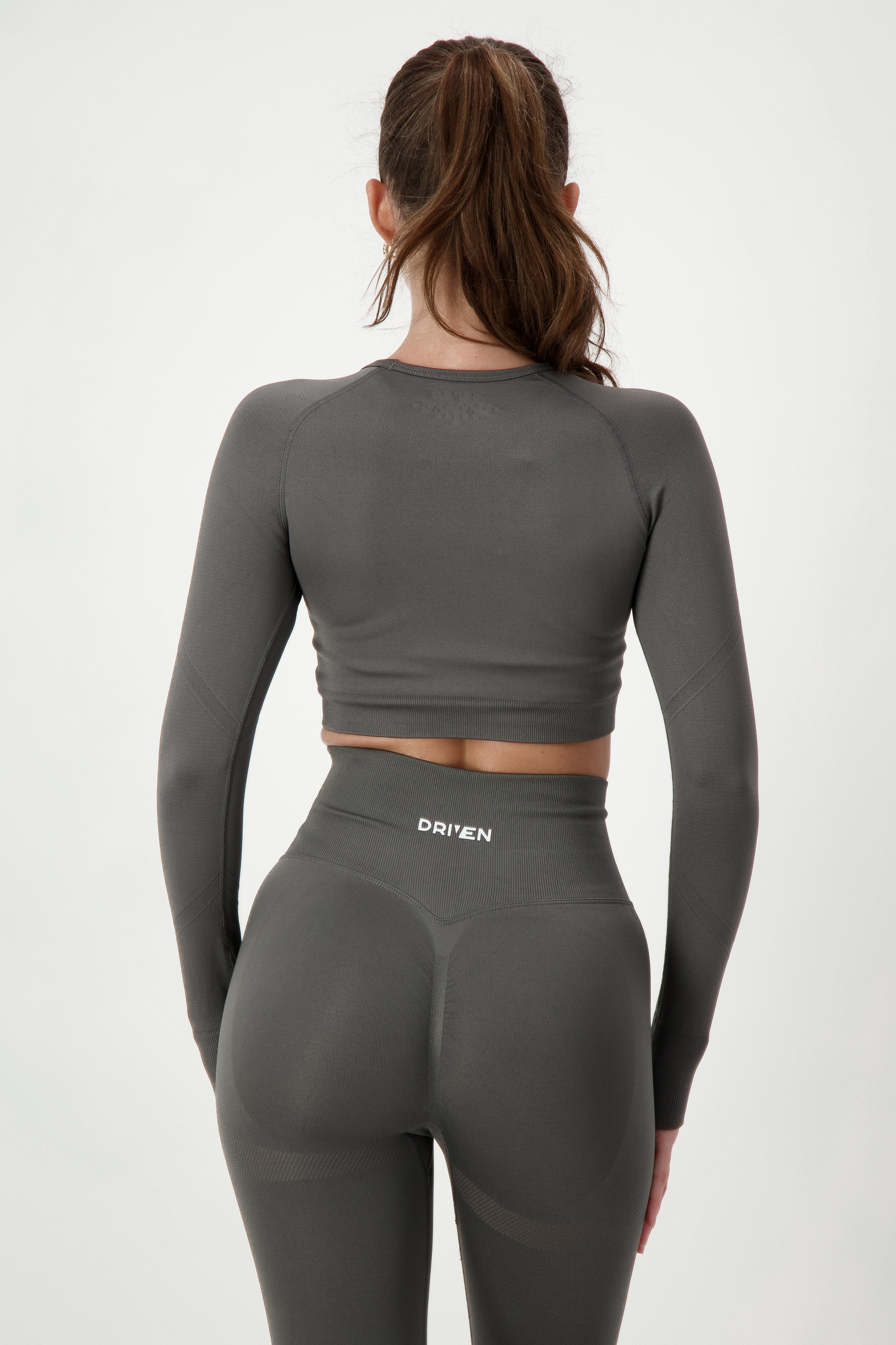 A woman with dark hair in a high ponytail is wearing a grey, form-fitting athletic jumpsuit and white sneakers. She has tattoos on her arm and is smiling while looking to the side. She stands against a plain white background.
