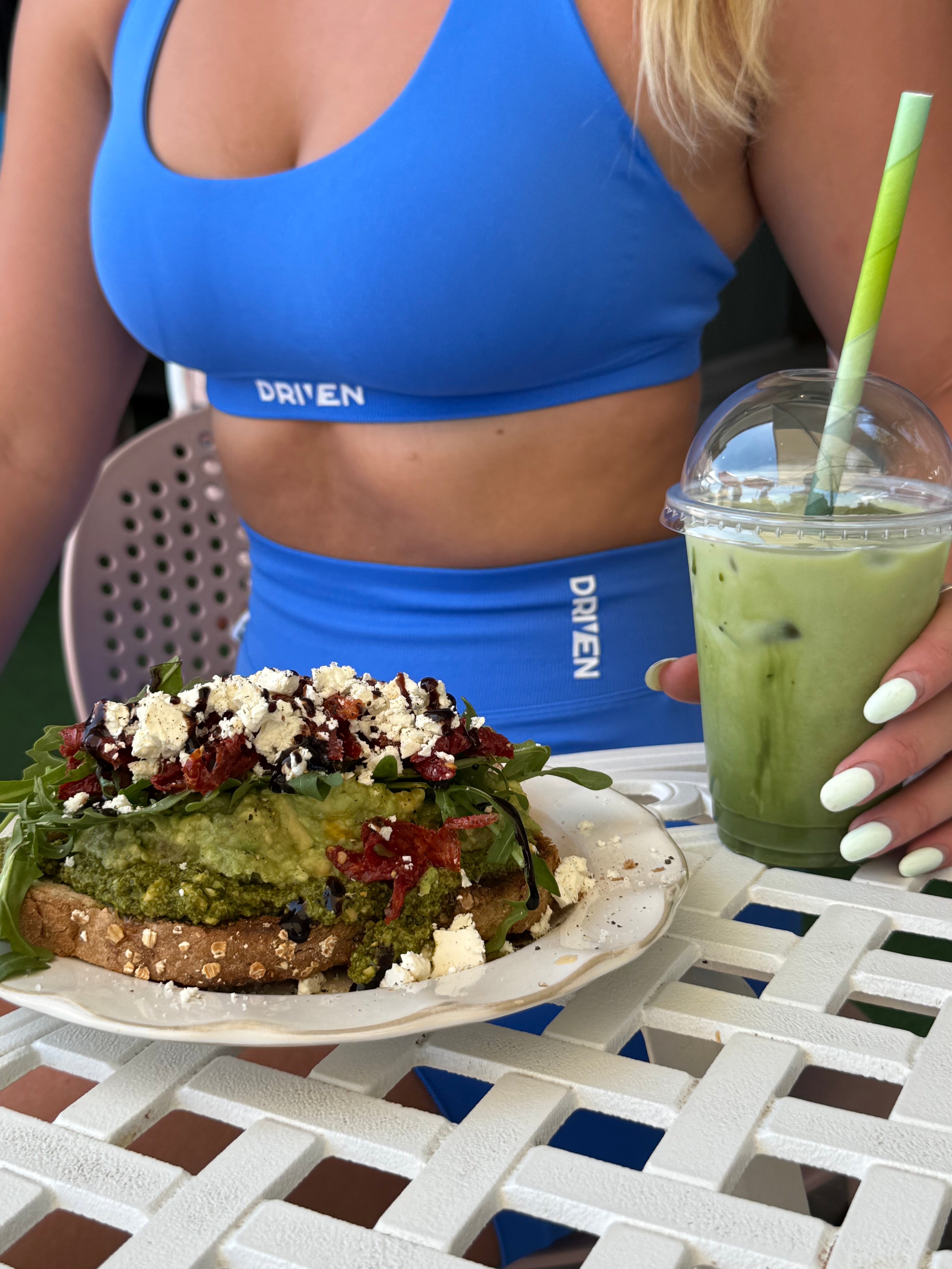 A woman with a ponytail is shown from the back. She is wearing a beige criss-cross sports bra and matching high-waist leggings which have the word "DRIVEN" printed on the waistband. She stands with her hands on her hips against a plain white background.