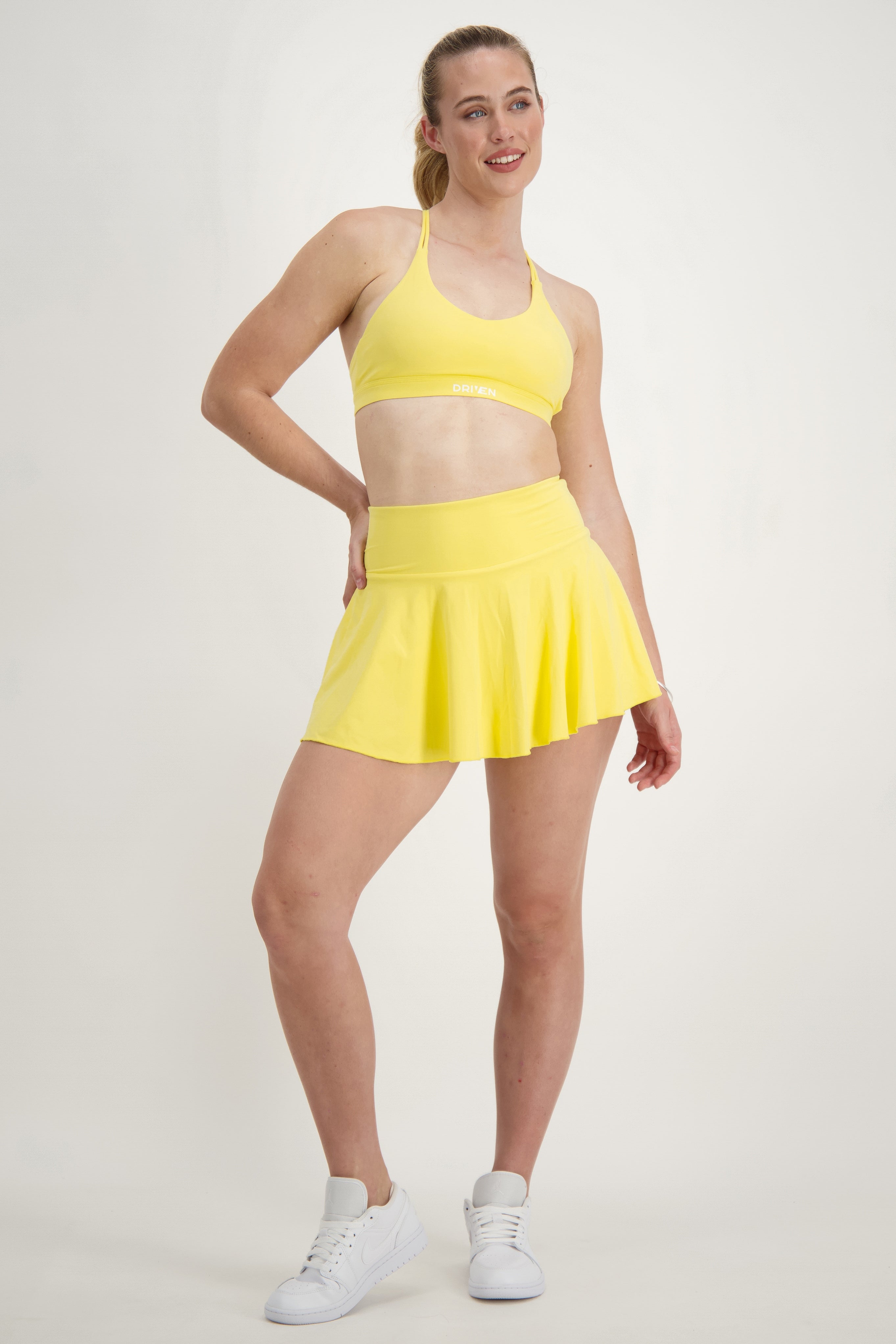 A woman with a ponytail is smiling at the camera. She is wearing a bright yellow outfit, consisting of a tank top and shorts. She is lifting part of her tank top with one hand, revealing a bit of her midriff. The background is plain white.