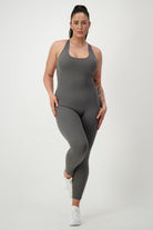 A person with long dark hair in a ponytail is wearing a form-fitting gray Classic Bodysuit Long from DRIVEN Gym Apparel and white sneakers. Dressed in stylish activewear, they stand against a plain white background, posing with one leg slightly bent and hands resting on their hips.