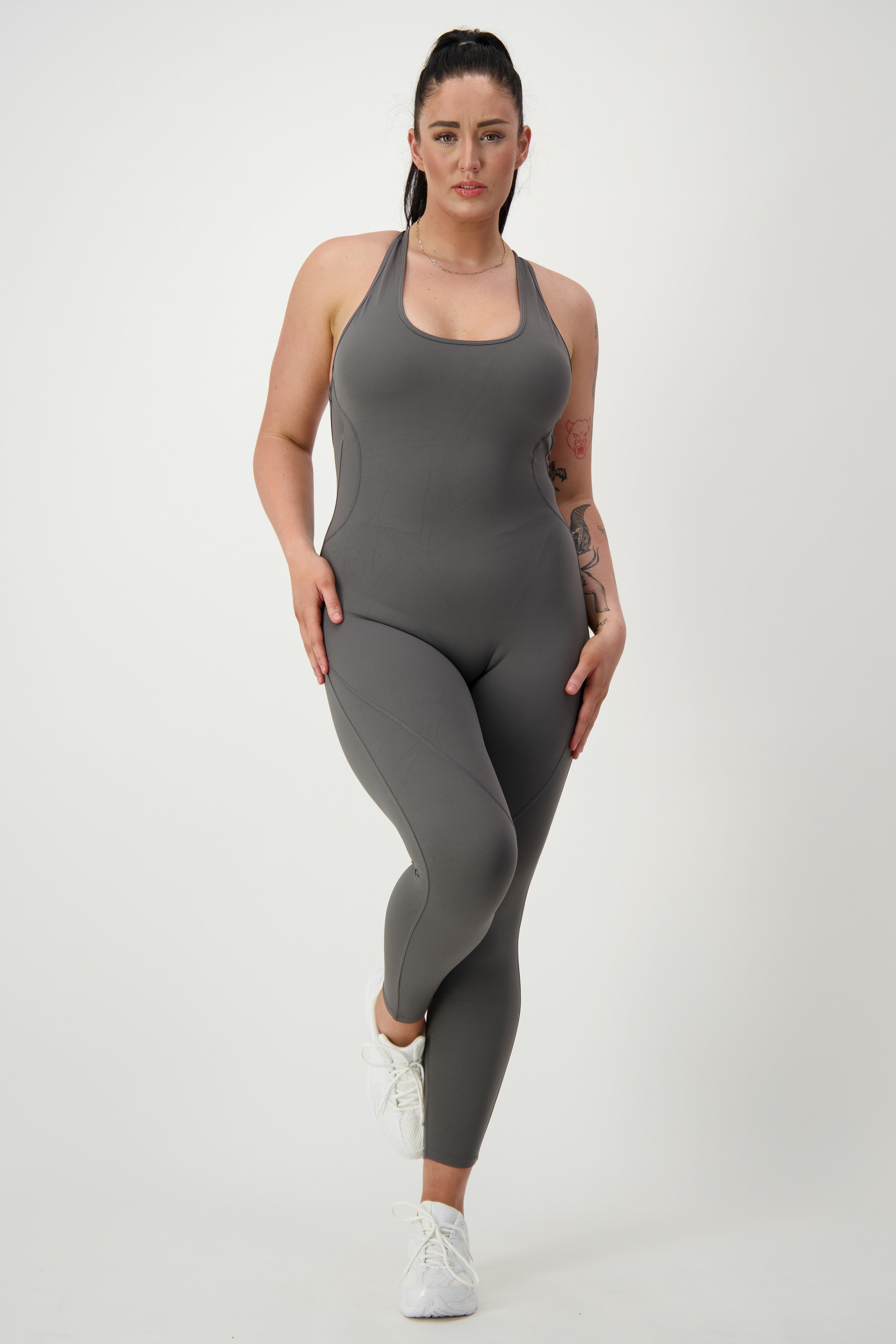 A person with long dark hair in a ponytail is wearing a form-fitting gray Classic Bodysuit Long from DRIVEN Gym Apparel and white sneakers. Dressed in stylish activewear, they stand against a plain white background, posing with one leg slightly bent and hands resting on their hips.