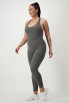 A woman with a high ponytail is wearing a form-fitting, grey **Classic Bodysuit Long** from **DRIVEN Gym Apparel** and white sneakers. She is walking and smiling, showcasing her tattoos. The chic athleisure look stands out against the plain white background.