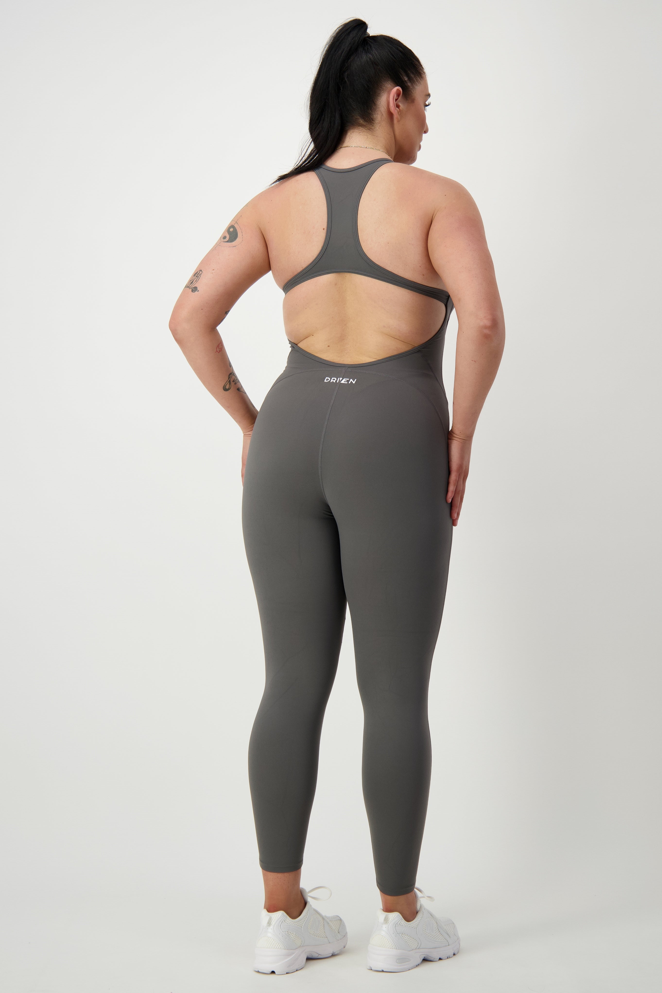 A person with long dark hair in a ponytail is standing against a plain background. They are wearing the grey Classic Bodysuit Long from DRIVEN Gym Apparel and white sneakers, looking slightly to the side with hands resting on their hips. Visible tattoos are on the person's arms.