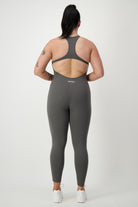 A person with a ponytail is standing with their back facing the camera, wearing a gray, sleeveless workout jumpsuit and white sneakers. The Classic Bodysuit Long (grey) has a racerback design and the brand name "DRIVEN Gym Apparel" is printed on the upper back. The background is plain white.