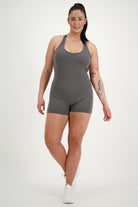 A woman with dark hair in a ponytail is standing against a plain white background, showcasing her grey, sleeveless Classic Bodysuit Short from DRIVEN Gym Apparel and white sneakers. Embracing athleisure style, she has tattoos on her left arm and is smiling while looking directly at the camera.