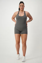A woman with dark hair in a ponytail is standing against a plain white background. She is wearing a grey Classic Bodysuit Short from DRIVEN Gym Apparel and white sneakers. She has several tattoos on her arms and is holding her hands behind her back.