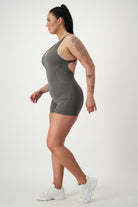 A person with a ponytail is walking to the left, wearing a form-fitting gray Classic Bodysuit Short (grey) by DRIVEN Gym Apparel and white sneakers. They have tattoos on their visible arm, showcasing stylish fitness apparel. The background is plain white.
