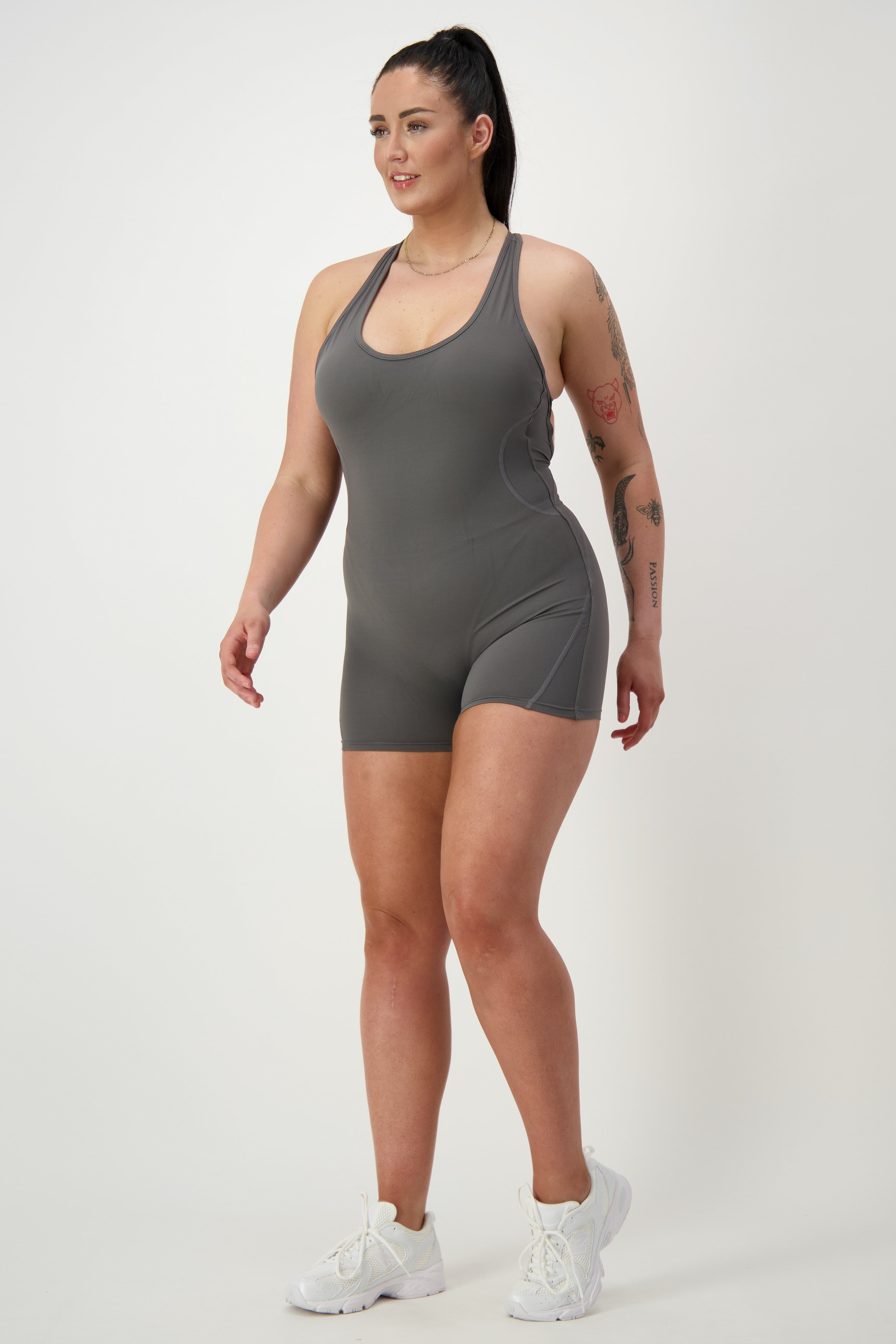 A woman with a ponytail is wearing a grey sleeveless Classic Bodysuit Short from DRIVEN Gym Apparel and white sneakers. She is standing against a plain white background, looking to the side, with visible tattoos on her arms.