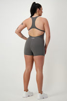 A person with long dark hair is standing facing away from the camera, wearing a DRIVEN Gym Apparel Classic Bodysuit Short (grey) and white sneakers. They have tattoos on their right arm and leg. The background is plain and light-colored.