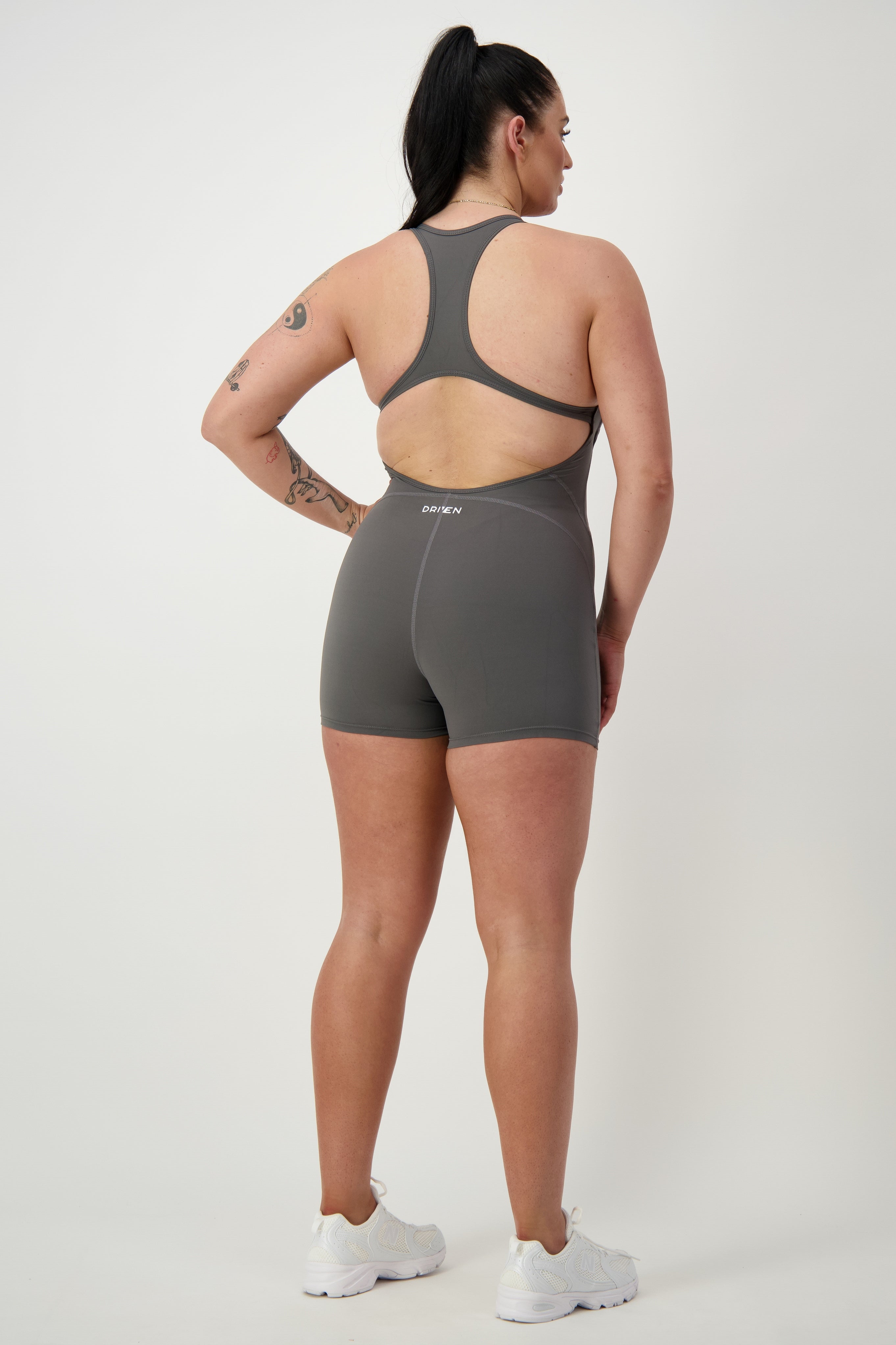 A person with long dark hair is standing facing away from the camera, wearing a DRIVEN Gym Apparel Classic Bodysuit Short (grey) and white sneakers. They have tattoos on their right arm and leg. The background is plain and light-colored.