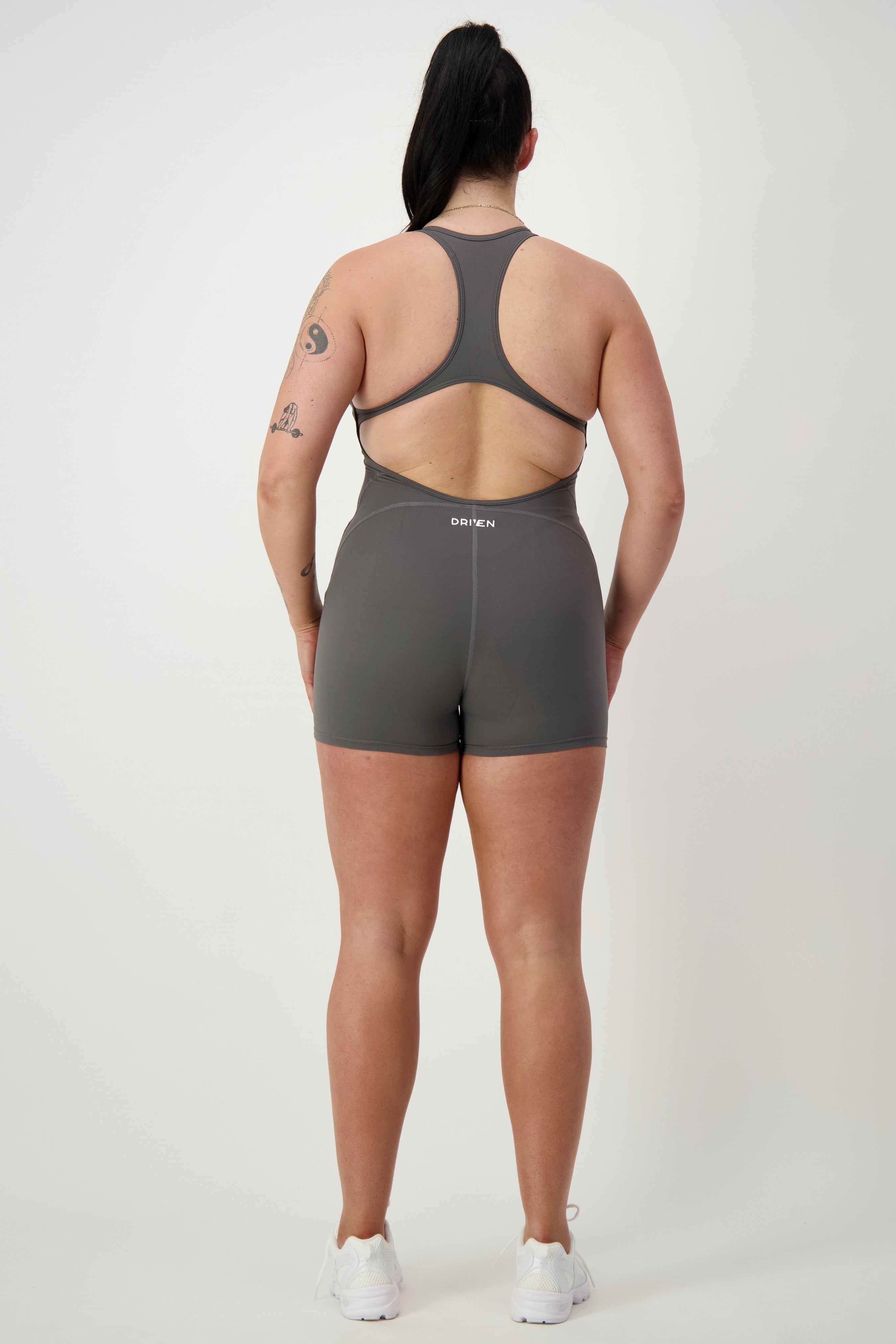 A person stands facing away from the camera, dressed in sleek gray activewear—a matching DRIVEN Gym Apparel Classic Bodysuit Short (grey). With their hair tied up and tattoos on their left arm, the bodysuit complements their white sneakers. The background is plain, highlighting their athleisure look.