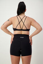 A person with long black hair in a ponytail is standing facing away, wearing black strappy gym wear including a sports bra and DRIVEN Gym Apparel Classic Midi Shorts (black) with the word "DRIVEN" on the waistband. The person has several tattoos on their arms and is posed against a plain white background.