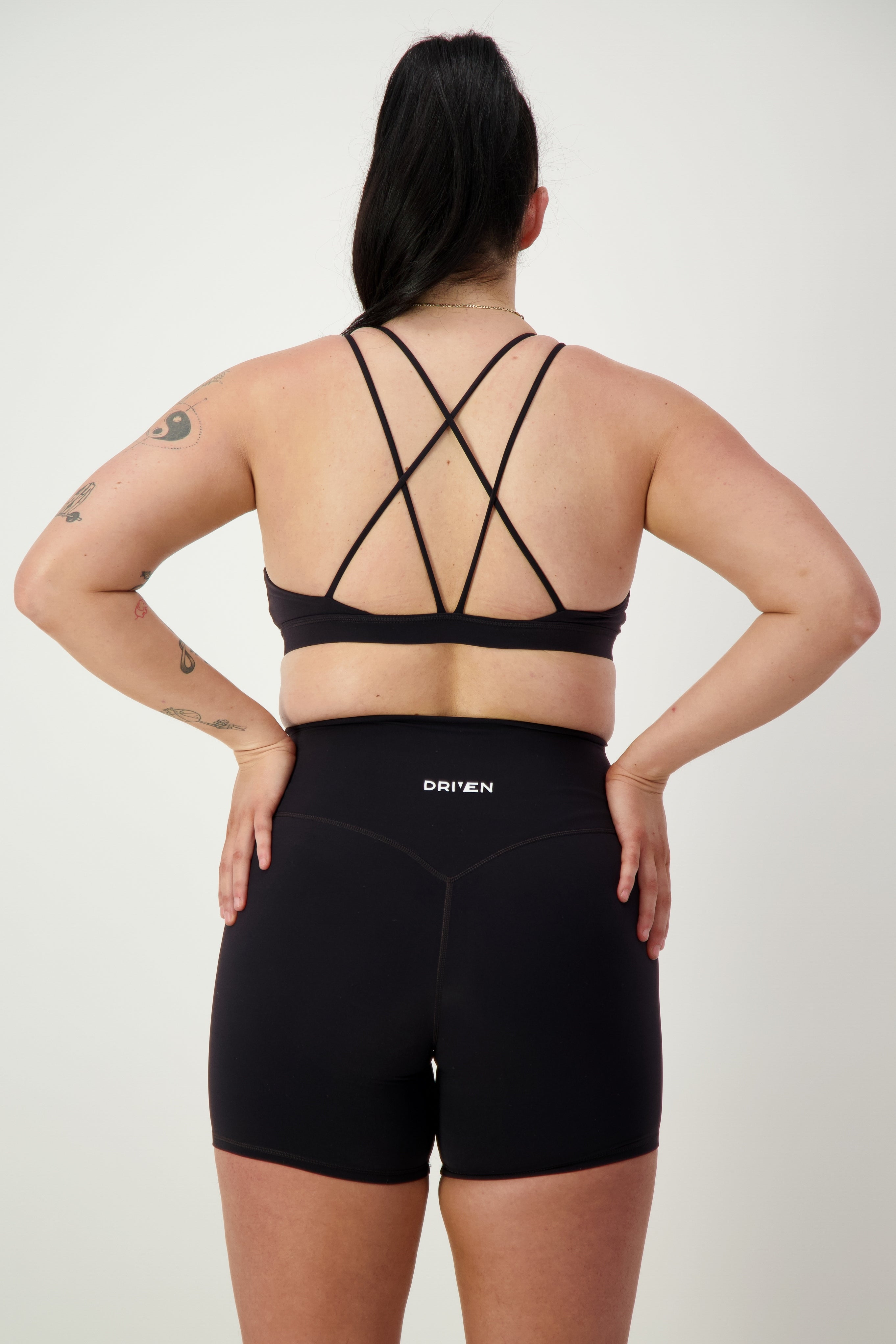 A person with long black hair in a ponytail is standing facing away, wearing black strappy gym wear including a sports bra and DRIVEN Gym Apparel Classic Midi Shorts (black) with the word "DRIVEN" on the waistband. The person has several tattoos on their arms and is posed against a plain white background.