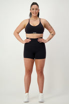 A woman in black gym wear stands confidently against a plain white background. She is wearing a sports bra and Classic Midi Shorts (black) by DRIVEN Gym Apparel, with white sneakers. Her hair is pulled back, and visible tattoos adorn her right arm. She is smiling and looking forward, embodying athleisure style perfectly.