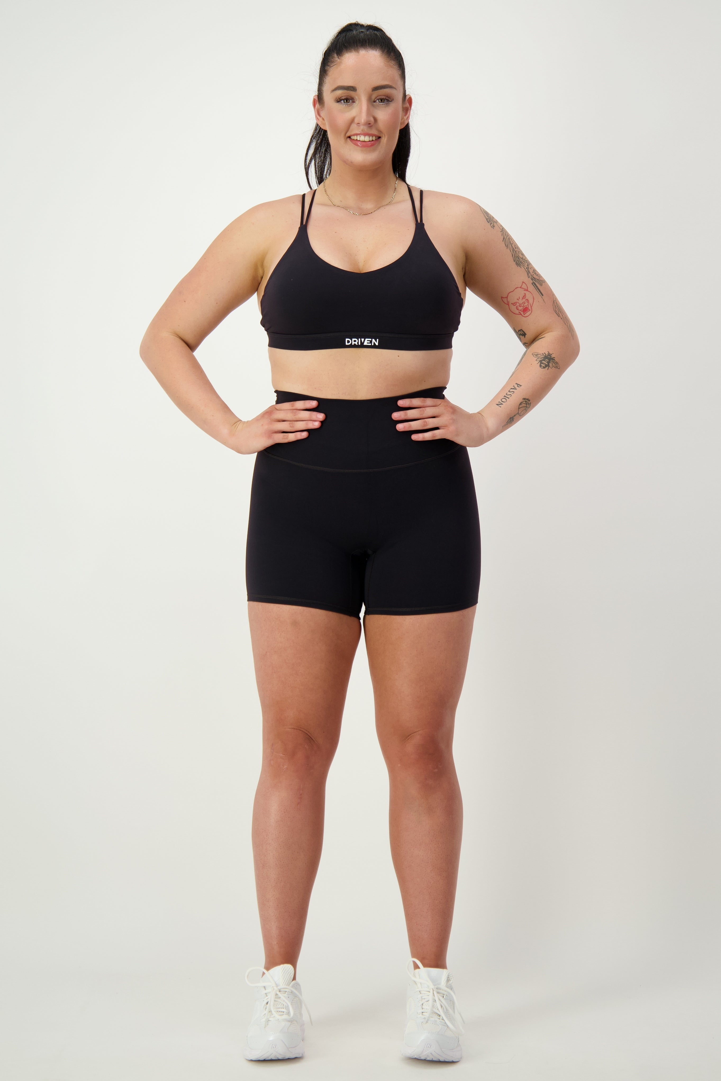 A woman in black gym wear stands confidently against a plain white background. She is wearing a sports bra and Classic Midi Shorts (black) by DRIVEN Gym Apparel, with white sneakers. Her hair is pulled back, and visible tattoos adorn her right arm. She is smiling and looking forward, embodying athleisure style perfectly.