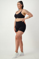 A woman stands against a plain background wearing black fitness apparel consisting of a sports bra and DRIVEN Gym Apparel's Classic Midi Shorts (black). She has tattoos on her left arm and is wearing white athletic shoes. Her dark hair is tied back in a high ponytail, and she is looking off to the side.