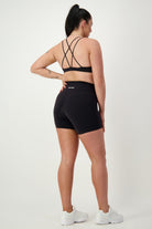 A woman stands with her back to the camera in stylish fitness apparel, featuring a black sports bra with crisscross straps and high-waisted Classic Midi Shorts (black) by DRIVEN Gym Apparel. Her black hair is tied up in a ponytail, and she completes the look with white sneakers. The background is plain white.