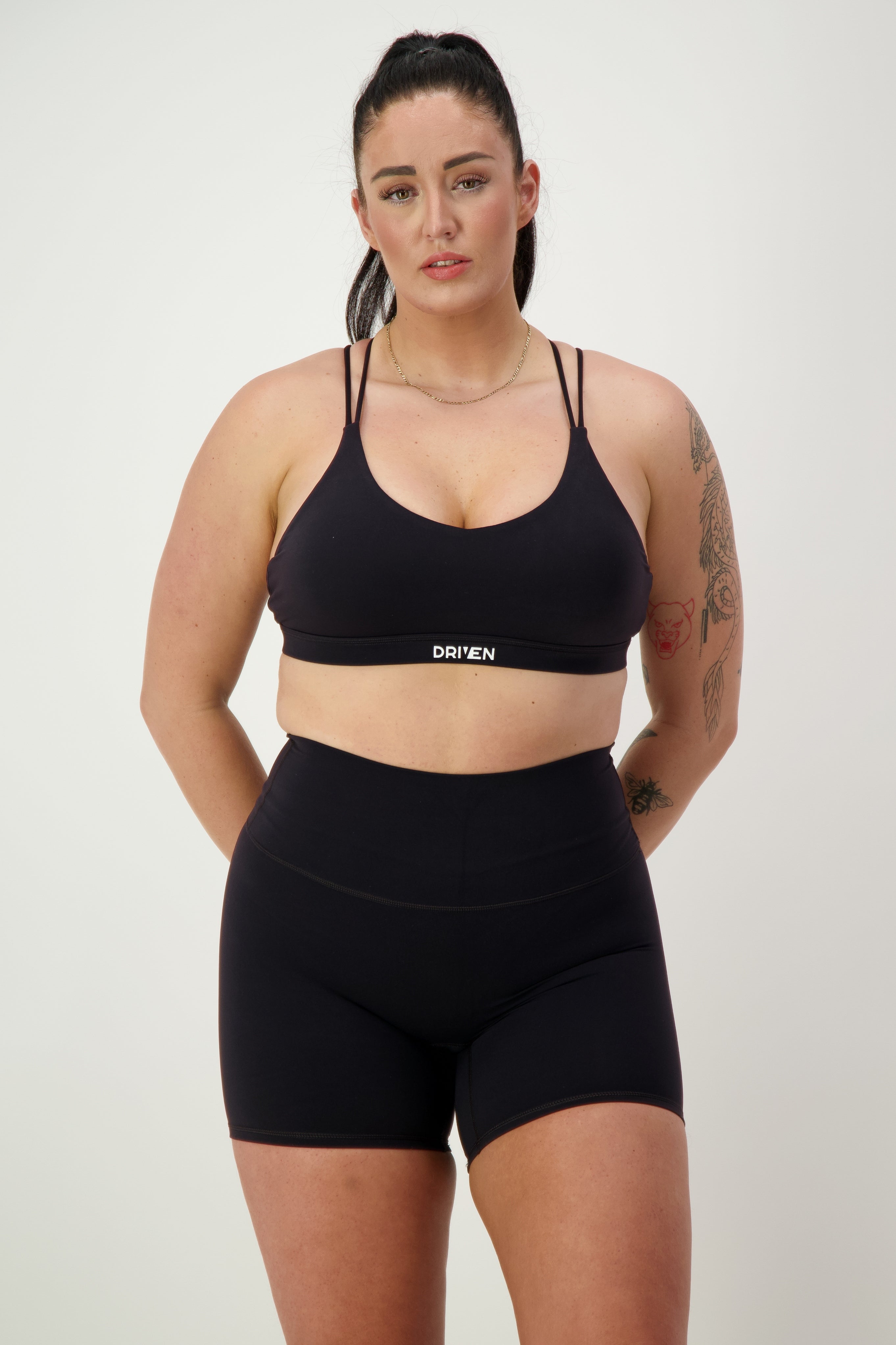 A woman with long dark hair is standing against a plain background, wearing black athleisure—a sleek sports bra and Classic Midi Shorts (black) from DRIVEN Gym Apparel. She has tattoos on her left arm and is looking directly at the camera with a neutral expression. The word "DRIVEN" is printed on her bra.