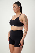 A woman with long dark hair in a high ponytail is wearing sleek black fitness apparel, including a sports bra and DRIVEN Gym Apparel's Classic Midi Shorts (black). She has tattoos on her left upper arm and forearm. Standing against a plain light background, she looks to the side with a focused expression.