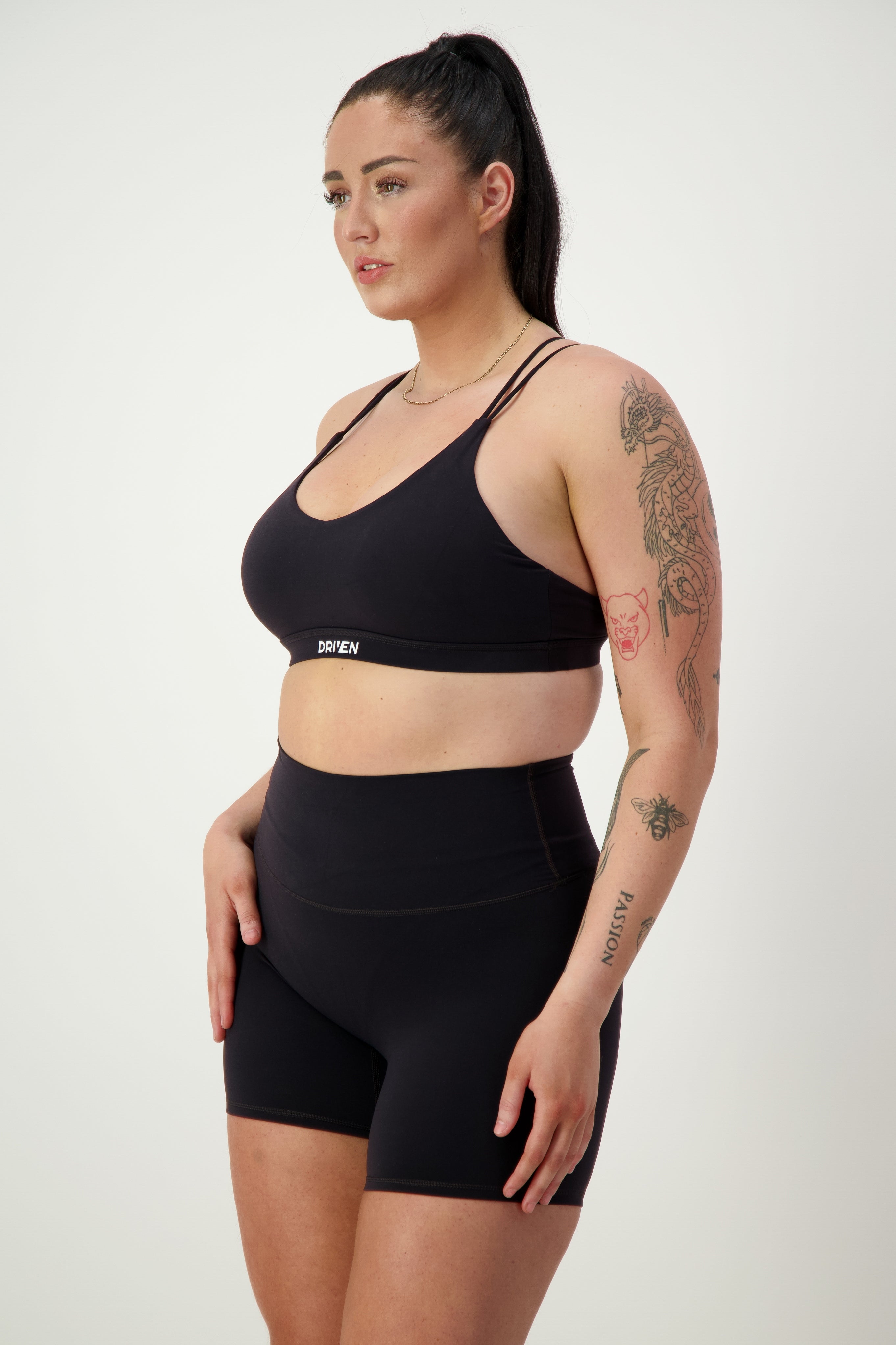 A woman with long dark hair in a high ponytail is wearing sleek black fitness apparel, including a sports bra and DRIVEN Gym Apparel's Classic Midi Shorts (black). She has tattoos on her left upper arm and forearm. Standing against a plain light background, she looks to the side with a focused expression.