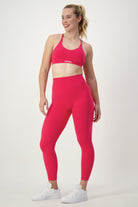 A woman is standing and smiling in a confident pose, wearing matching bright pink activewear—a sports bra and DRIVEN Gym Apparel's Classic Full Length Tights (cherry). She has her hair tied back in a ponytail and is also wearing white sneakers. The background is a plain, light-colored studio backdrop.