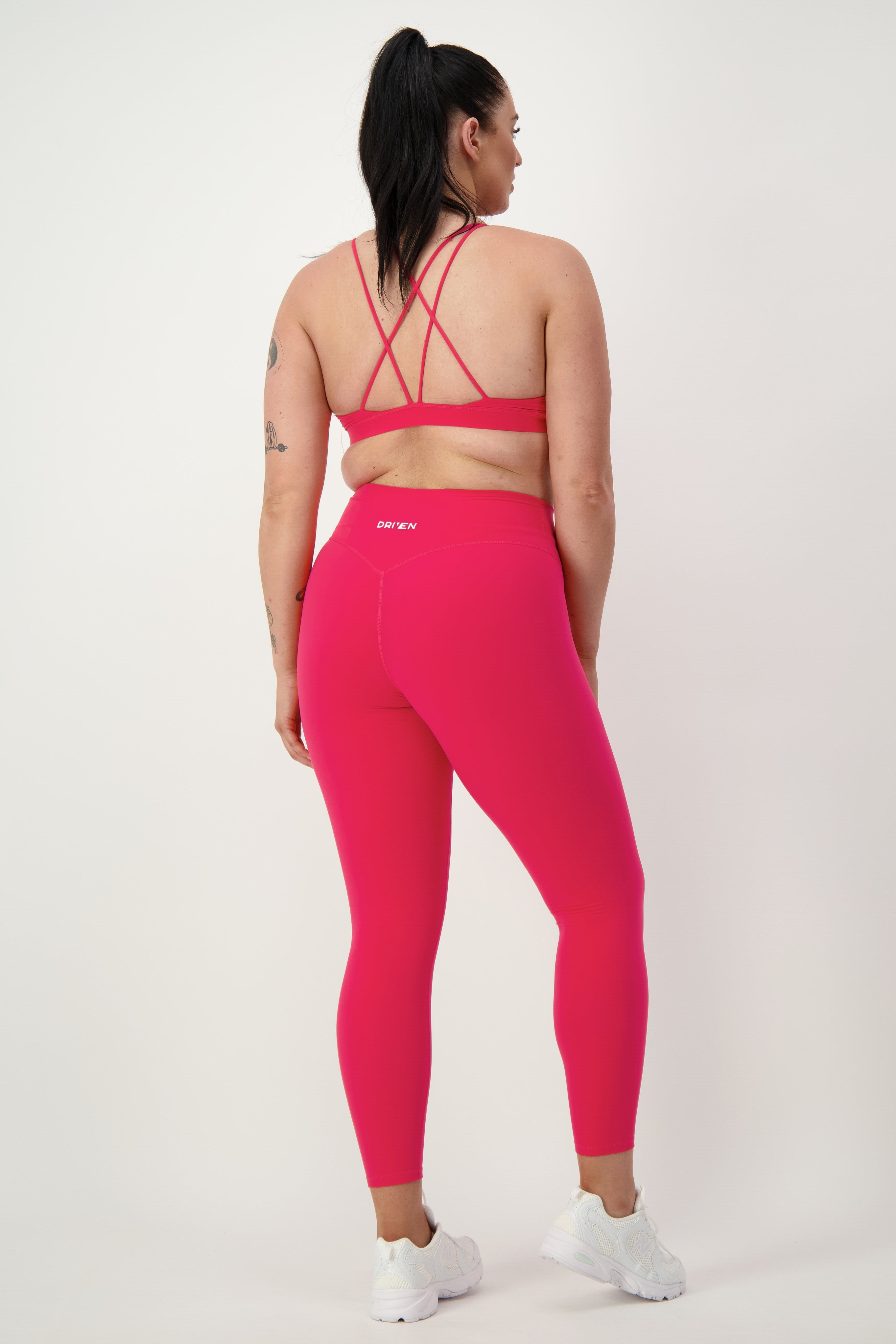 A woman is standing facing away from the camera, showcasing her gym wear. She is wearing a matching bright pink sports bra and DRIVEN Gym Apparel's Classic Full Length Tights (cherry) with white sneakers. She has a ponytail and visible tattoos on her arm. The background is plain white, highlighting the stylish athleisure outfit perfect for fitness aficionados.