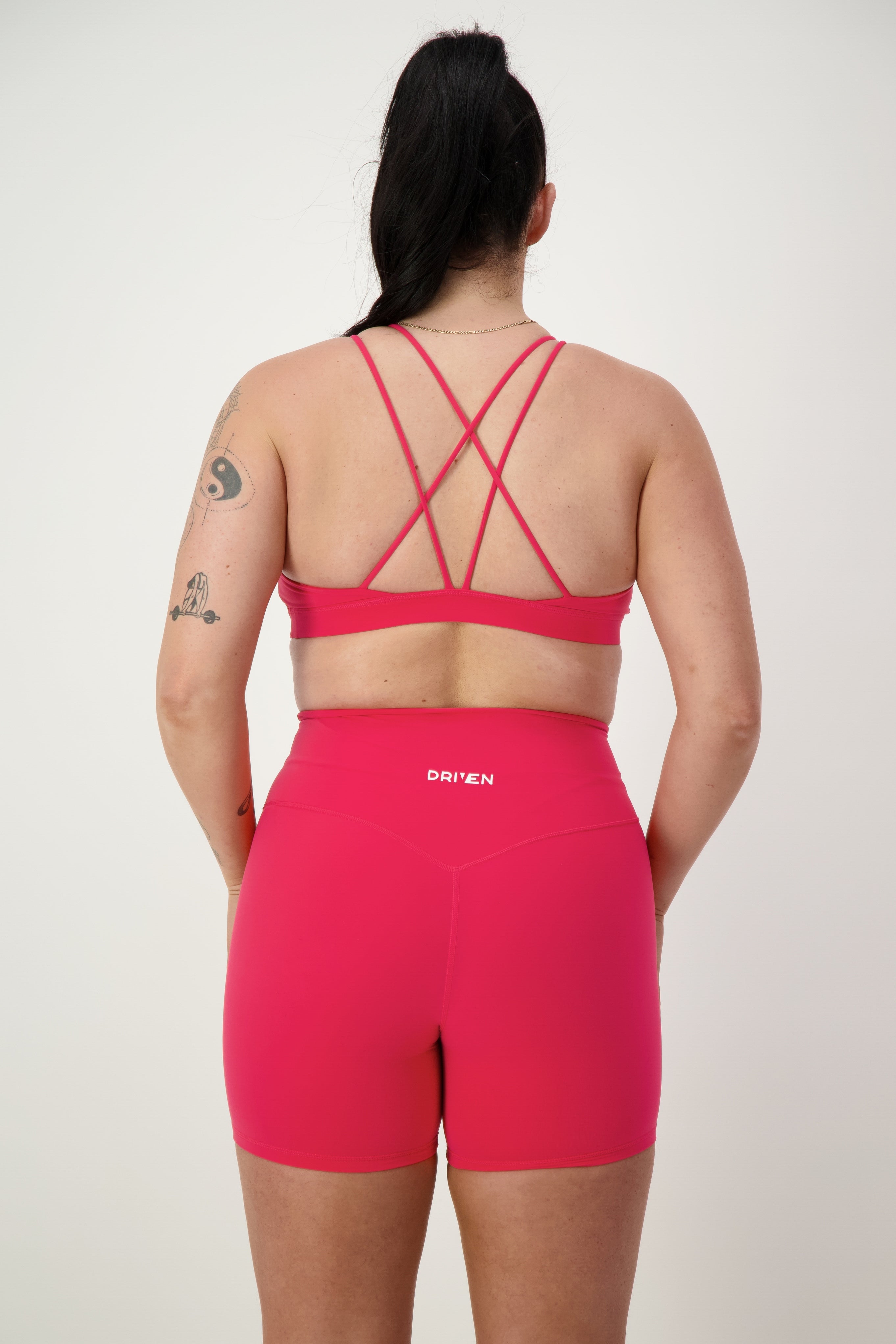 A person is seen from behind wearing a DRIVEN Gym Apparel Classic Strappy Back Bra (cherry) with matching high-waisted shorts with the word "DRIVEN" printed on the waistband. Their stylish activewear showcases a tattoo on their upper right arm, and their hair is tied up in a high ponytail.