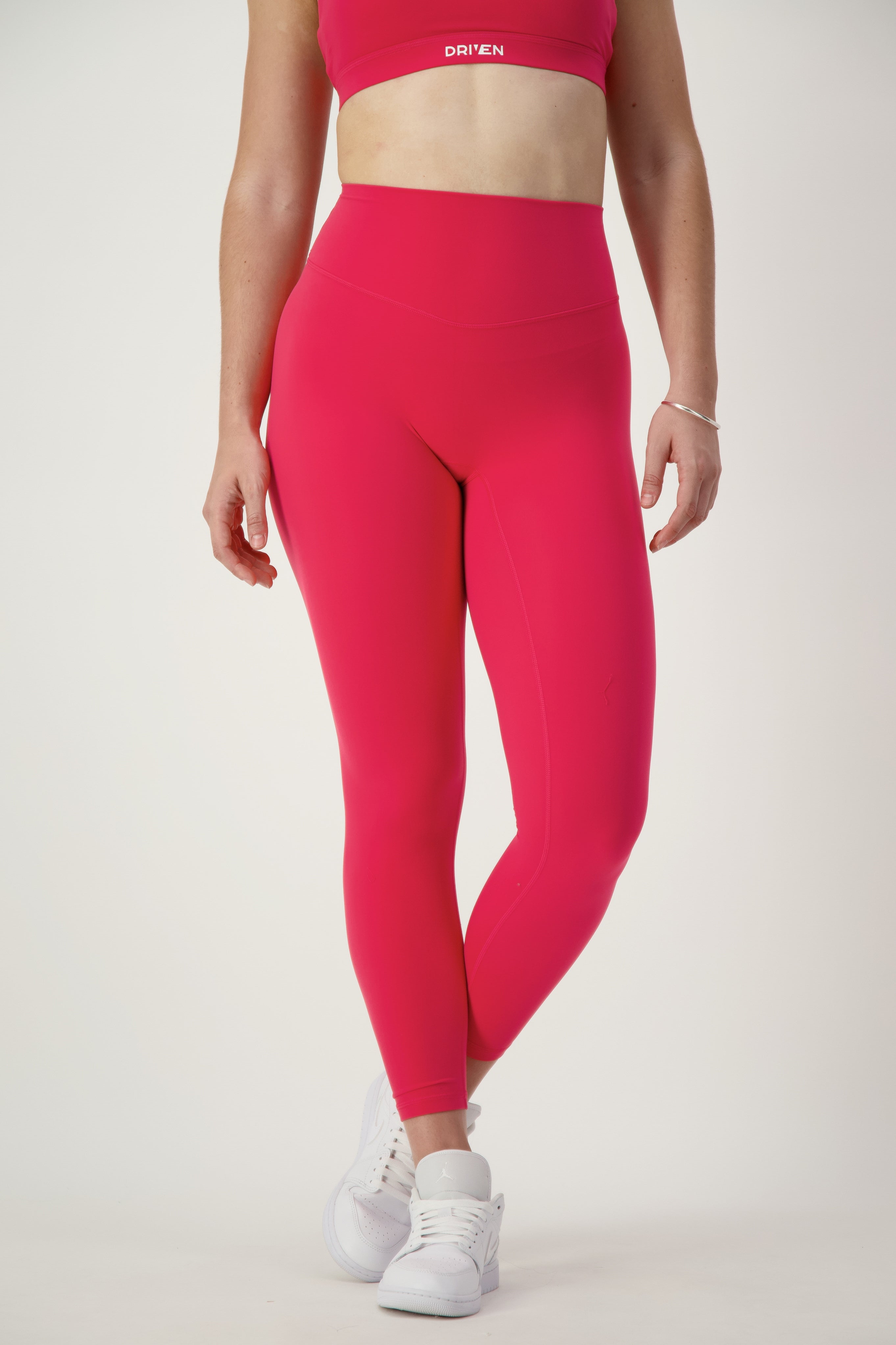A person is wearing a red sports bra and matching Classic Full Length Tights (cherry), epitomizing modern activewear. They have white sneakers on and a bracelet on their left wrist. Standing against a plain white background, their lower face, arms, and legs are visible.