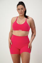 A woman with long dark hair in a high ponytail is wearing pink fitness apparel, including a Classic Strappy Back Bra (cherry) and matching high-waisted shorts. She stands against a simple white background, showcasing several tattoos on her right arm and left thigh. The sports bra features the word "DRIVEN Gym Apparel" in white text.