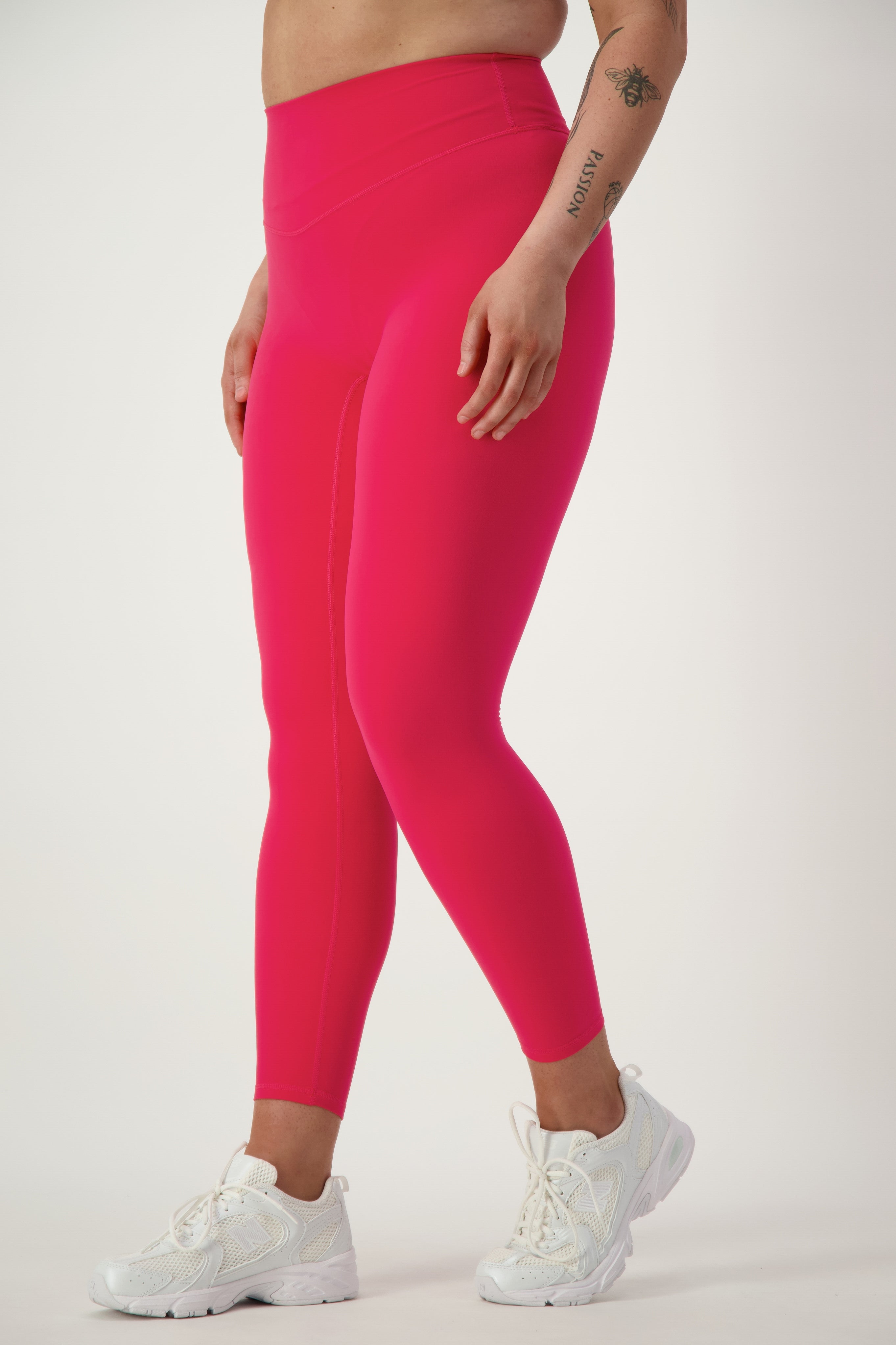 A person wearing bright pink Classic Full Length Tights (cherry) by DRIVEN Gym Apparel and white sneakers, exuding athleisure vibes, stands against a plain white background. One arm has visible tattoos, and their hand rests by their side. Their upper body is not visible in the image.