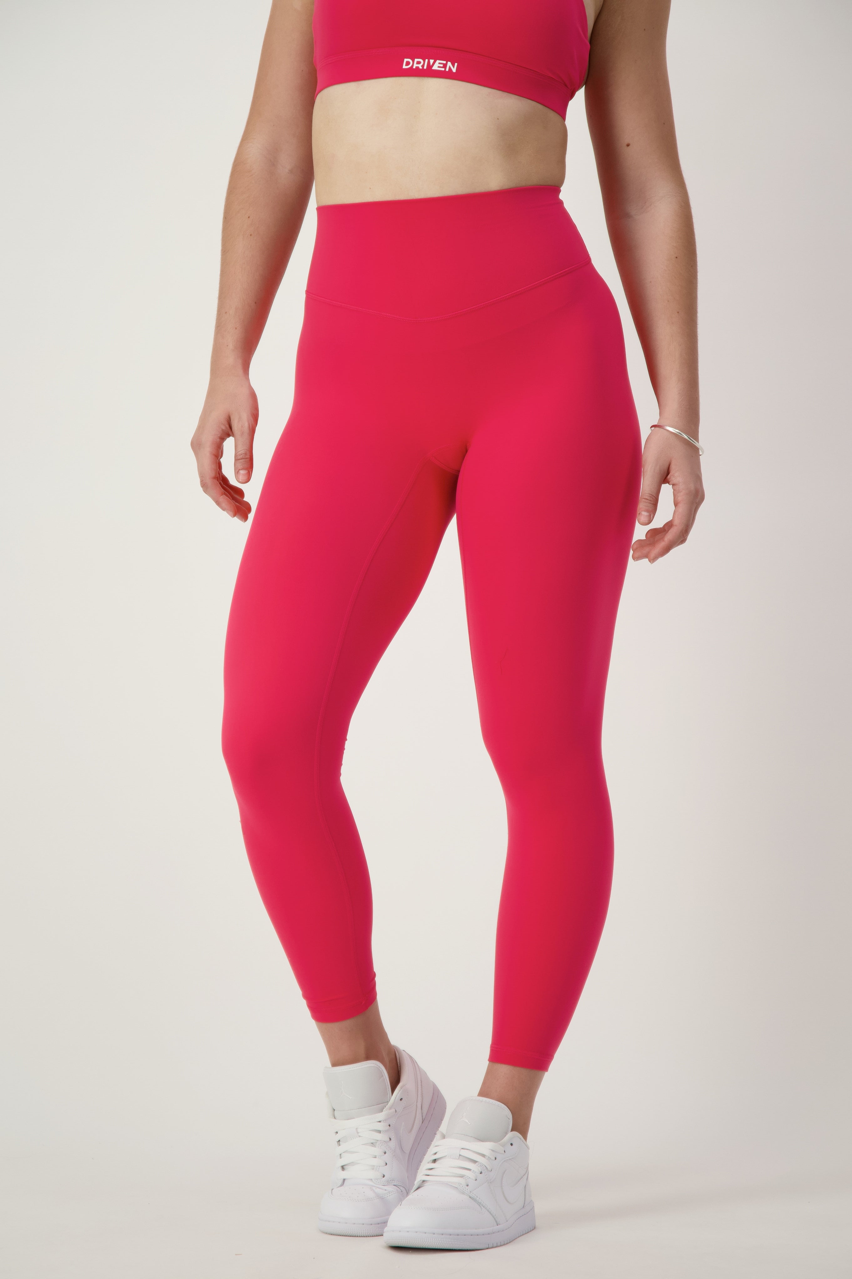 A person wearing matching red fitness apparel, including a sports bra and DRIVEN Gym Apparel's Classic Full Length Tights (cherry), stands against a plain white background. Their outfit is complemented with white athletic shoes, and a minimal bracelet is noticeable on their left wrist.