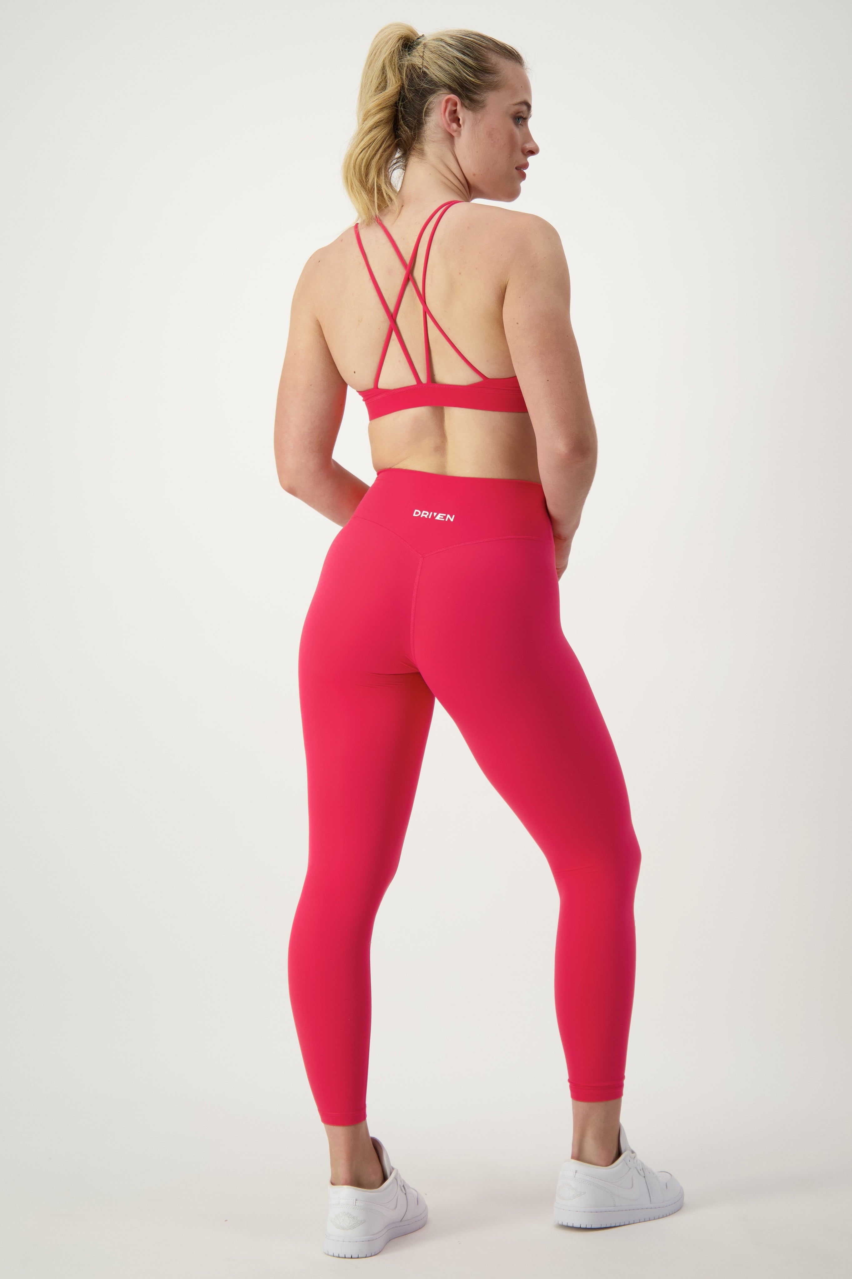 A woman in a bright pink sports bra with crisscross straps and matching DRIVEN Gym Apparel Classic Full Length Tights (cherry), epitomizing athleisure, stands facing away. She has a ponytail and is wearing white sneakers. The background is plain white.