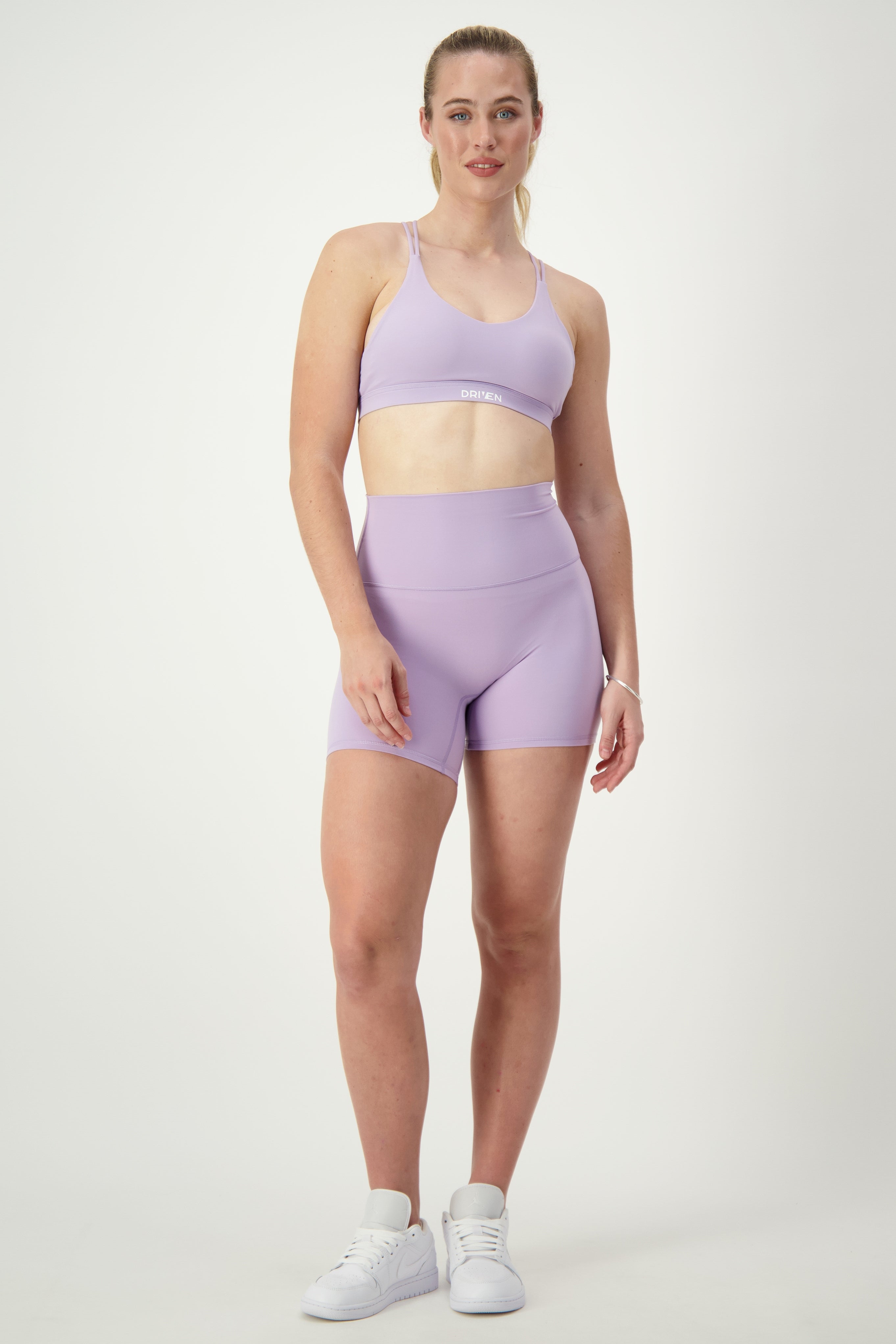 A woman stands against a plain background, showcasing her athleisure look with a Classic Strappy Back Bra (iris) by DRIVEN Gym Apparel and matching high-waisted shorts. She has her hair pulled back and is wearing white sneakers. With a neutral expression, her hands are relaxed by her sides, perfectly embodying modern gym wear.
