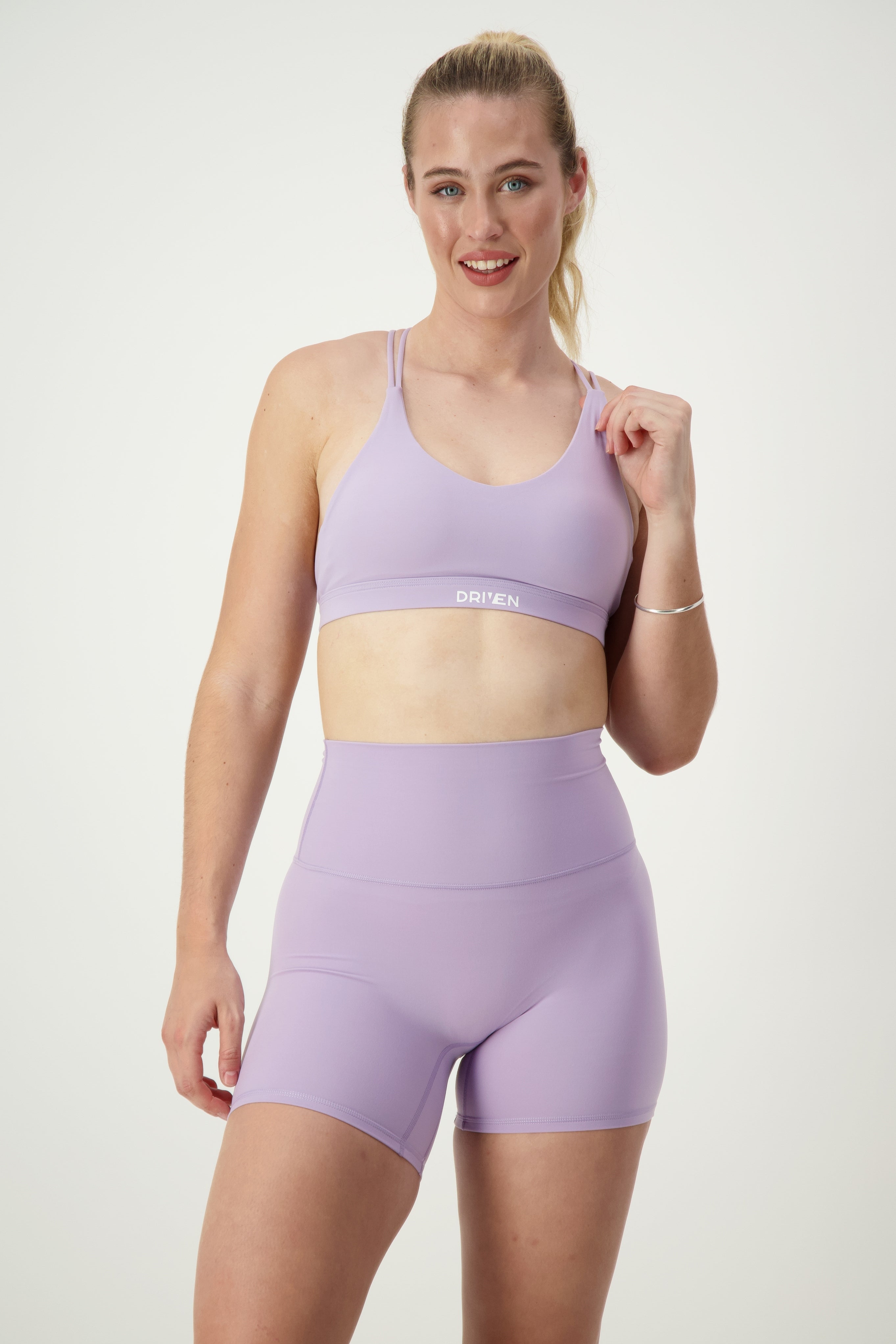 A person with long hair in a ponytail is smiling at the camera, showcasing their matching light purple fitness apparel, including a DRIVEN Gym Apparel Classic Strappy Back Bra (iris) and high-waisted athletic shorts. The background is plain white.
