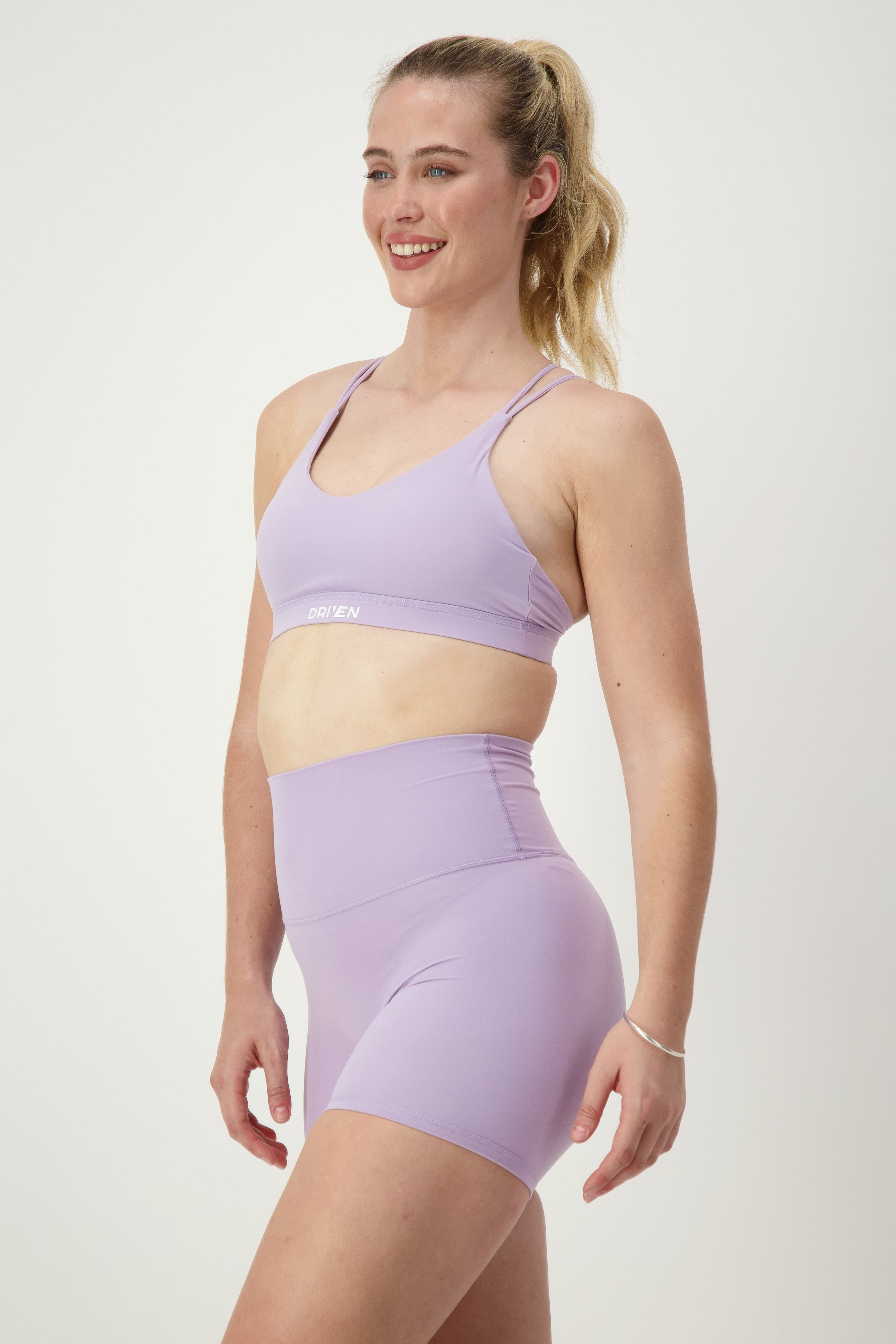A person with long blonde hair tied in a ponytail is smiling and wearing matching purple athleisure—a Classic Strappy Back Bra (iris) by DRIVEN Gym Apparel and high-waisted shorts. They are standing against a plain white background.