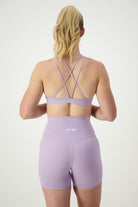 A woman with blonde hair in a ponytail is seen from the back wearing light purple fitness apparel: the Classic Strappy Back Bra (iris) and matching high-waisted shorts. The word "DRIVEN" is printed on the back of the shorts. The background is plain white.
