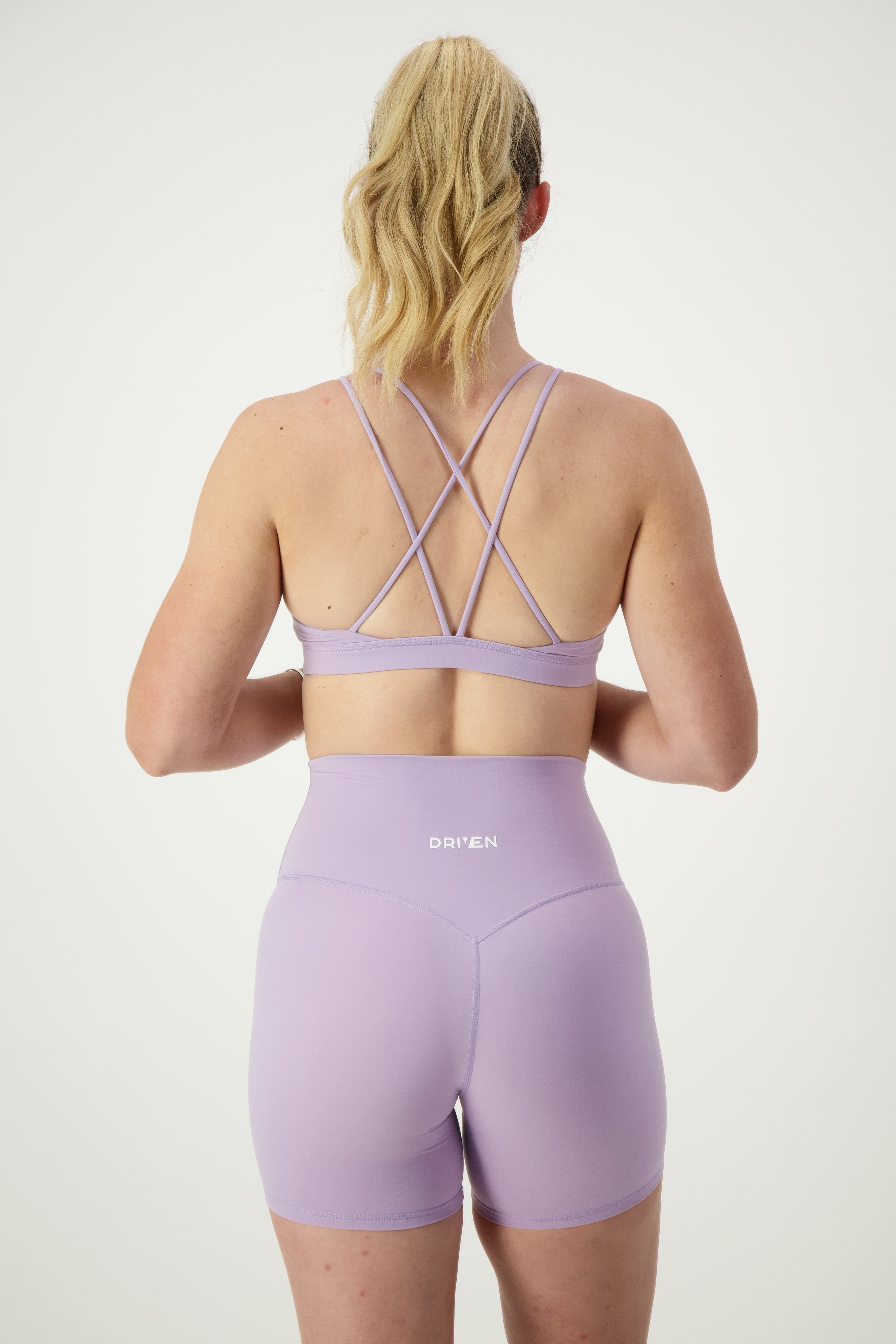 A woman with blonde hair in a ponytail is seen from the back wearing light purple fitness apparel: the Classic Strappy Back Bra (iris) and matching high-waisted shorts. The word "DRIVEN" is printed on the back of the shorts. The background is plain white.