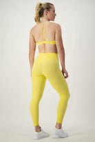 A woman with a ponytail is seen from the back, wearing yellow activewear: DRIVEN Gym Apparel's Classic Full Length Tights (lemon) and a matching sports bra with crisscross straps. She stands in a brightly lit room on a light grey background, wearing white sneakers.