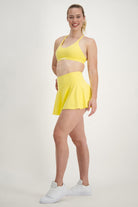 A woman stands against a plain white background, smiling at the camera in her vibrant activewear. She is wearing a bright yellow sports bra, matching yellow Classic Mini Skort (lemon) from DRIVEN Gym Apparel, and white sneakers. Her blonde hair is tied back in a ponytail, and she stands with one leg slightly in front of the other.