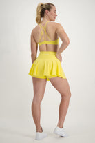 A woman stands facing away from the camera, showcasing vibrant athleisure in a bright yellow sports bra with criss-cross straps and a matching Classic Mini Skort (lemon) by DRIVEN Gym Apparel. Her hair is in a ponytail, and she's sporting white sneakers, all set against a plain white background.