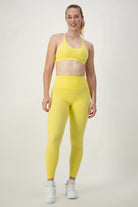 A woman stands against a plain background, smiling. She is wearing vibrant yellow activewear—a sports bra and matching high-waisted Classic Full Length Tights (lemon) by DRIVEN Gym Apparel, along with white athletic shoes. Her hair is pulled back into a ponytail. With a confident posture and her left hand resting by her side, she radiates fitness apparel style.