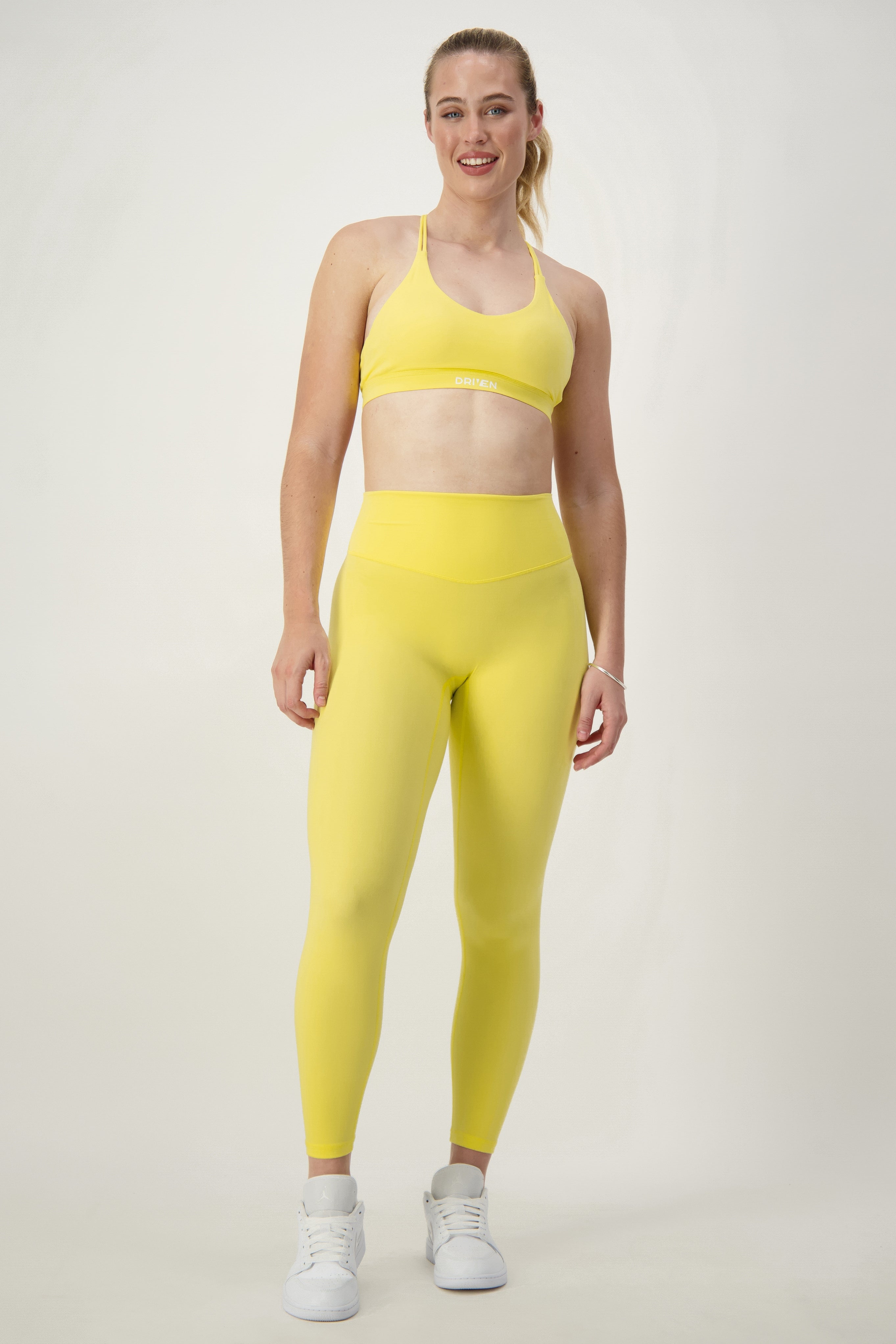 A woman stands against a plain background, smiling. She is wearing vibrant yellow activewear—a sports bra and matching high-waisted Classic Full Length Tights (lemon) by DRIVEN Gym Apparel, along with white athletic shoes. Her hair is pulled back into a ponytail. With a confident posture and her left hand resting by her side, she radiates fitness apparel style.