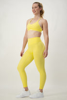 A woman stands against a plain background, smiling and looking to her left. She is dressed in bright yellow activewear—a sports bra and matching DRIVEN Gym Apparel Classic Full Length Tights (lemon)—paired with white sneakers. Her hair is tied back in a ponytail, and she wears a bracelet on her right wrist.