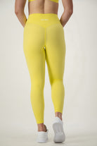 A person is standing with their back to the camera, wearing DRIVEN Gym Apparel's Classic Full Length Tights (lemon) and white sneakers. Shown from the waist down, the focus is on the tights. The background is plain and light-colored.