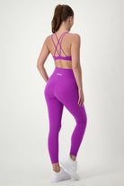 A woman stands facing away from the camera, wearing the Classic Strappy Back Bra (purple) from DRIVEN Gym Apparel with matching high-waisted leggings from our latest activewear collection. She has white sneakers on, her hair tied back in a ponytail, all perfectly showcasing this stylish fitness apparel against a plain white background.