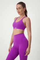 A person with long brown hair tied back stands against a plain background wearing matching purple activewear, consisting of a Classic Strappy Back Bra (purple) by DRIVEN Gym Apparel and high-waisted leggings. They are looking towards the camera with a neutral expression.