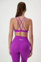 A person with long hair in a ponytail is shown from the back, wearing a DRIVEN Gym Apparel Classic Strappy Back Bra (purple) and matching high-waisted leggings. The word "DRIVEN" is printed on the waistband of the leggings, embodying stylish athleisure. The background is plain white.