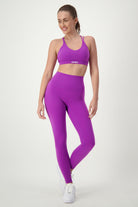 A woman is standing and smiling in athleisure, consisting of a purple Classic Strappy Back Bra from DRIVEN Gym Apparel and matching high-waisted leggings. Her hair is tied back, and she’s wearing white sneakers. The background is plain white.