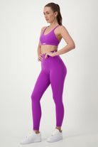 A woman stands in a side pose wearing matching purple fitness apparel, consisting of a Classic Strappy Back Bra (purple) from DRIVEN Gym Apparel and high-waisted leggings. She has her hair tied back in a ponytail and is sporting white sneakers. The background is plain white.
