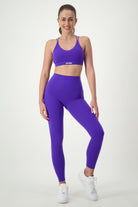 A woman is standing against a plain white background, wearing matching purple fitness apparel consisting of a sports bra and Classic Full Length Tights (sapphire). She's also wearing white sneakers and has her hair tied back in a ponytail. The brand logo "DRIVEN Gym Apparel" is visible on her sports bra.